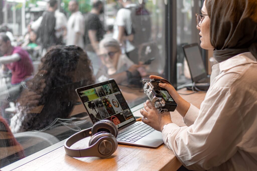 A woman in a hijab using a laptop and camera in a cafe setting, illustrating remote work.
